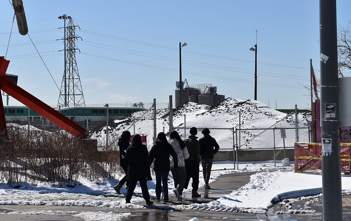A group of students walk down a sidewalk in front of a statue in downtown Toronto on an urban planning themed scavenger hunt.